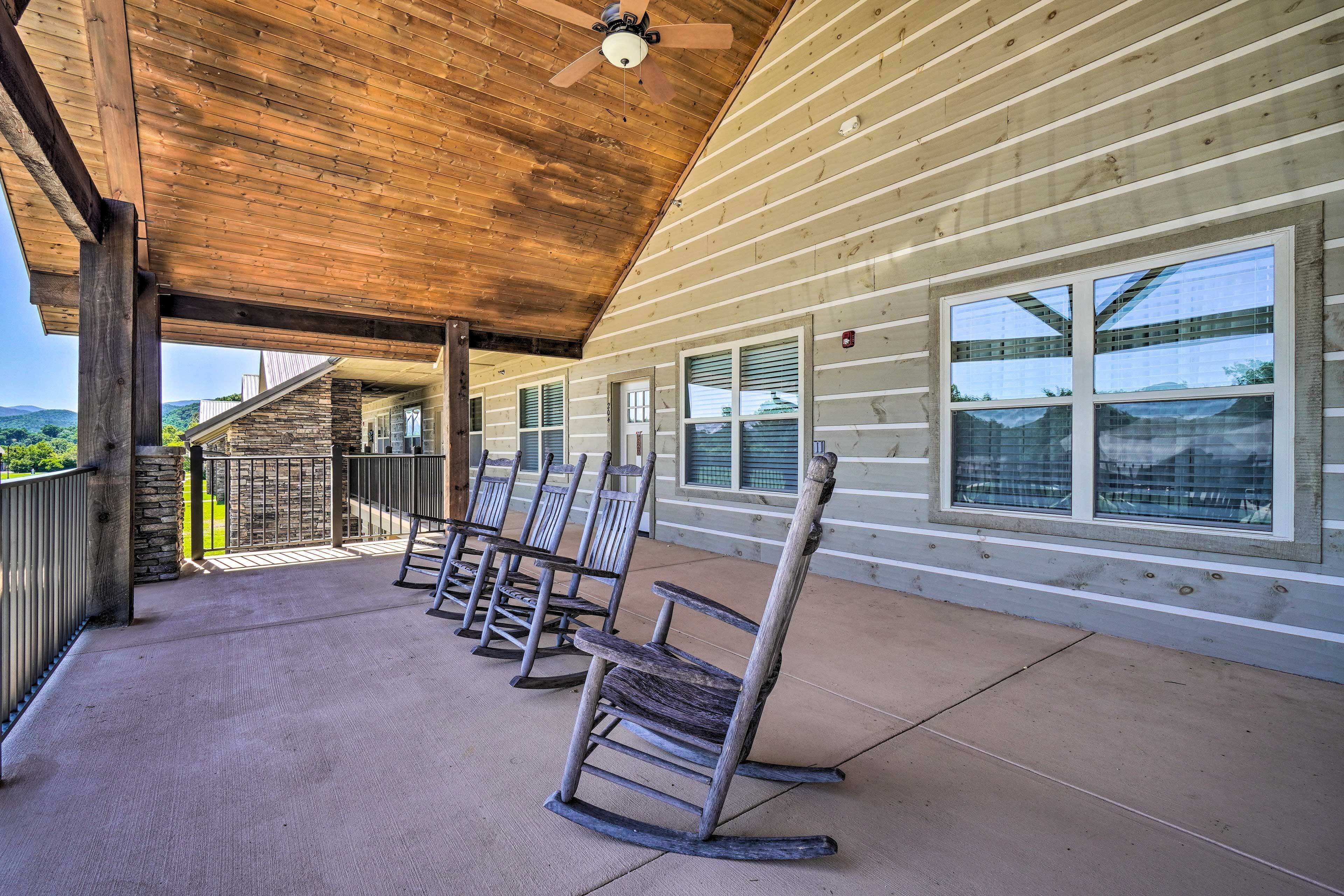 A covered outdoor area with wooden rocking chairs, a ceiling fan, and large windows overlooking a scenic landscape.