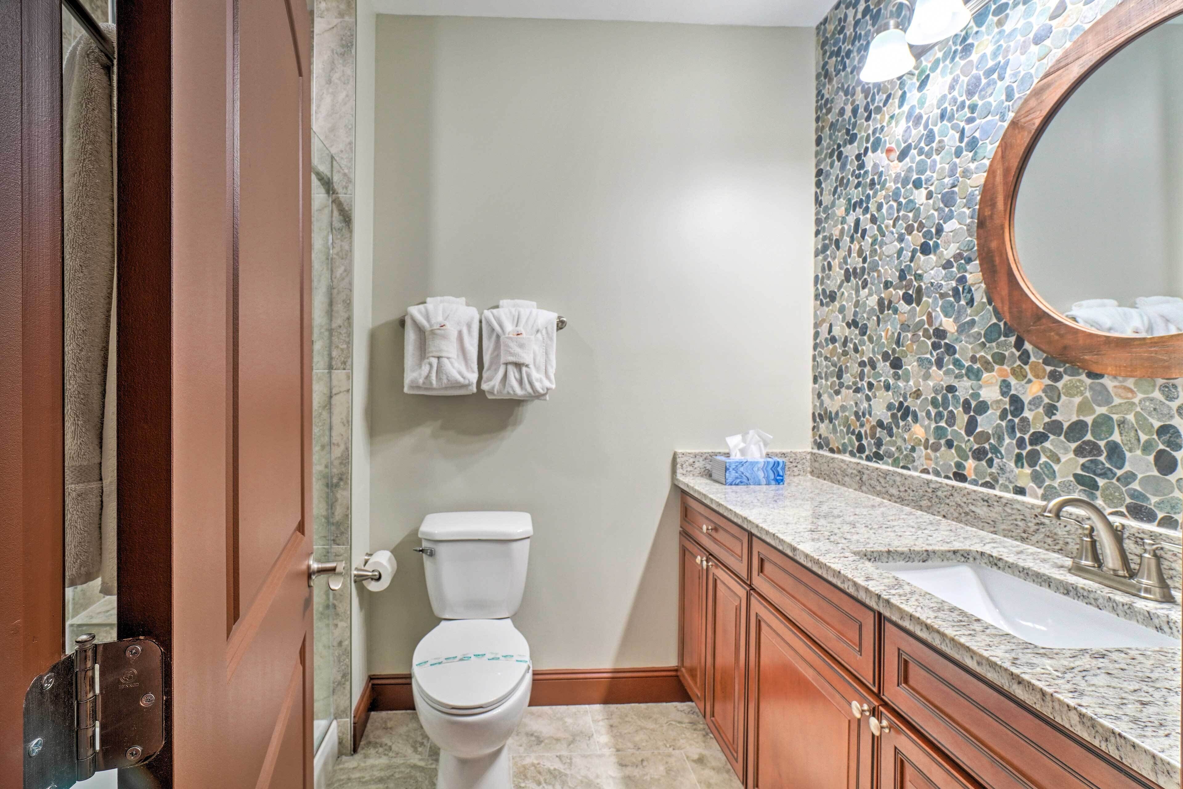 A bathroom with a toilet, wooden cabinets, a granite countertop, a sink, and a mosaic wall.