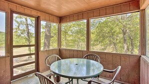 A covered patio with a round table and chairs, large windows with a view of trees, and wooden walls.