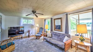 A living room with a ceiling fan, a flat-screen TV, a sofa, a chair, a lamp, and a window with curtains. - Cozy Ishpeming Cottage w/ Lake & Park Views! (Ishpeming)