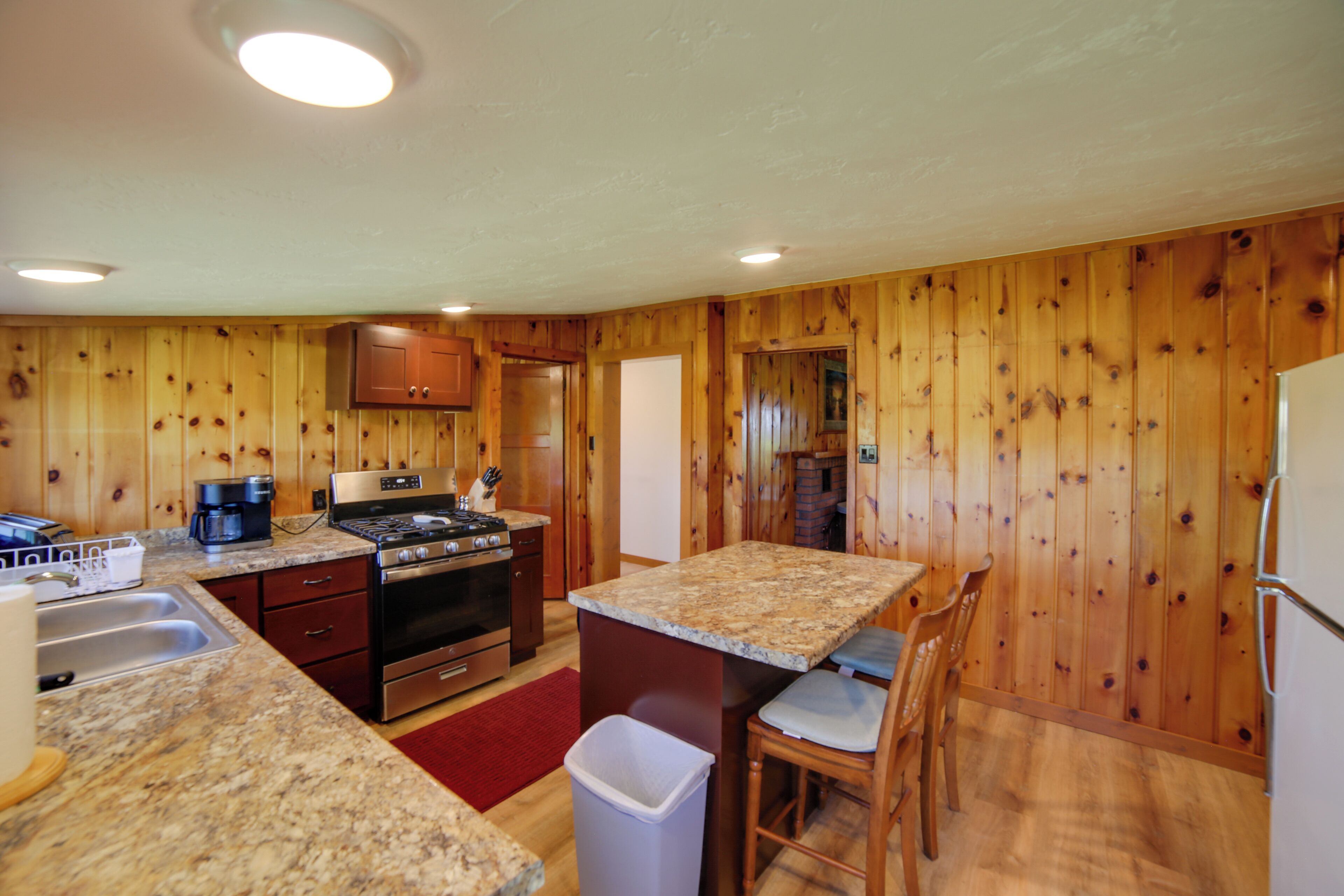 A compact kitchen with wooden paneling, a granite countertop, stainless steel appliances, and a small dining area with a chair.