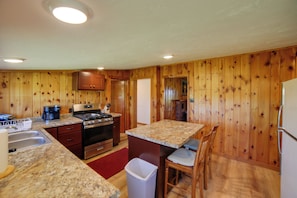 A compact kitchen with wooden paneling, a granite countertop, stainless steel appliances, and a small dining area with a chair. - 600' of Green Bay Frontage & Dock: Cozy Cottage (Oconto)