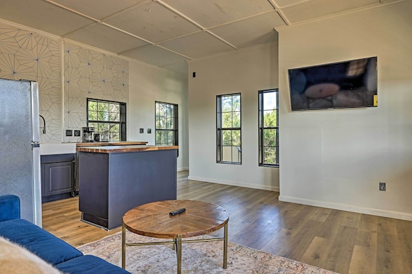 Amplia sala de estar con cocina, ventanas grandes y televisor montado.