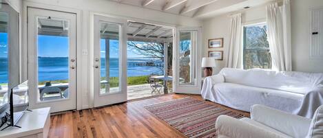 A spacious room with a wooden floor, white furniture, and large glass doors leading to a deck with a view of the ocean.