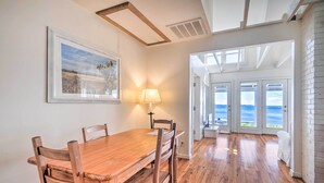 Un comedor con una mesa y sillas de madera, un cuadro en la pared y vista al océano a través de amplias puertas.