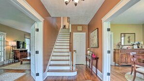 A staircase leading to a second floor, a dining area with a table and chairs, and a hallway with a door and a small table with items on it. - Elegant Charles Town Home: Grill, Walk Dtwn! (Charles Town)