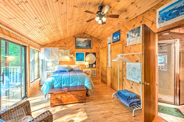 A cozy wooden cabin room with a bed, a wooden chest, a wicker chair, and a glass door leading to a balcony. - Dreamy Bucksport Hideaway on Freshwater Pond! (Bucksport)