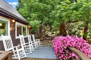 A wooden deck with white chairs and a blue patterned rug, surrounded by lush greenery and pink flowers. - Cozy Ferndale Studio on 20-acre Working Farm! (Ferndale)