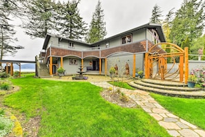 A two-story house with a covered patio, stone pathway, and a small garden with a fountain. - Custom Guemes Home w/ Spectacular Water Views! (Anacortes)