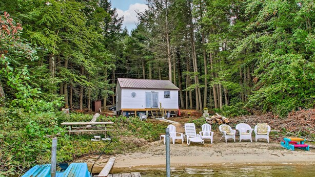 Una cabaña con un muelle, rodeada de un bosque frondoso y una playa arenosa con sillas de descanso.