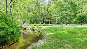 A rustic cabin with a wooden deck, surrounded by lush greenery and a small stream.