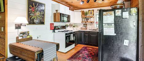 A kitchen with wooden cabinets, a black refrigerator, a stove, and a microwave. There is a dining table with a patterned tablecloth and two stools. A rug is on the floor, and a painting hangs on the wall.