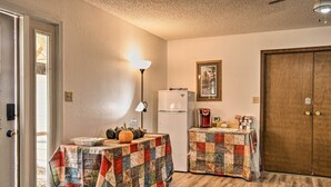 A compact kitchen with two tables covered in quilts, a refrigerator, a coffee maker, and a door with a coat hanging on it. - Highplaines Haven Apt - Fish, Hunt & Relax! (Long Lake)