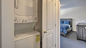 A laundry area with a front-loading washer and dryer, a door leading to a bedroom with a bed and a nightstand. - Glen Cove Home: Walk to Boardwalk + Dining! (Glen Cove)