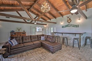 A spacious living area with a large brown leather sectional sofa, a wooden ceiling with exposed beams, a chandelier, and a rustic brick floor. - Theriot 'camp Outdoor Dreaming' Retreat w/ Dock! (Theriot)