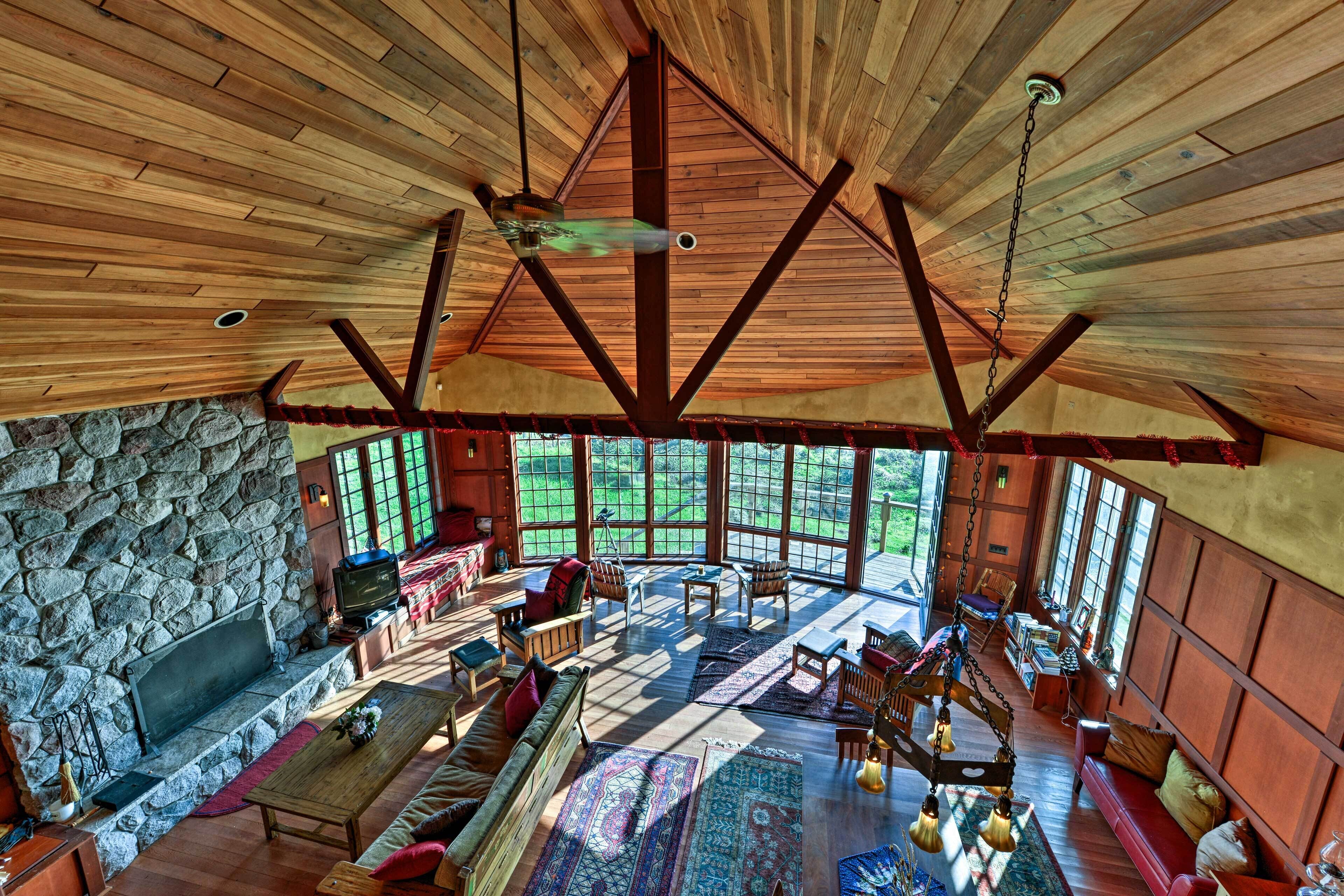 A spacious living area with a stone fireplace, wooden ceiling beams, and large windows offering a view of the outdoors.