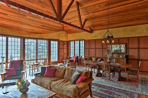 A spacious living room with wooden furniture, large windows, and a dining area. - Forested Gold Beach House w/ Panoramic Ocean Views (Gold Beach)