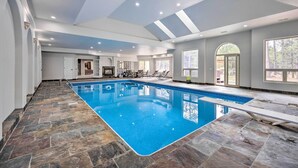 Uma grande piscina interior com pavimento de pedra, teto com iluminação embutida e área de estar com cadeiras e mesa.