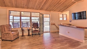 Un amplio salón con techo de madera, grandes puertas correderas de vidrio que dan a una terraza exterior, una barra de madera y un televisor de pantalla plana montado en la pared.