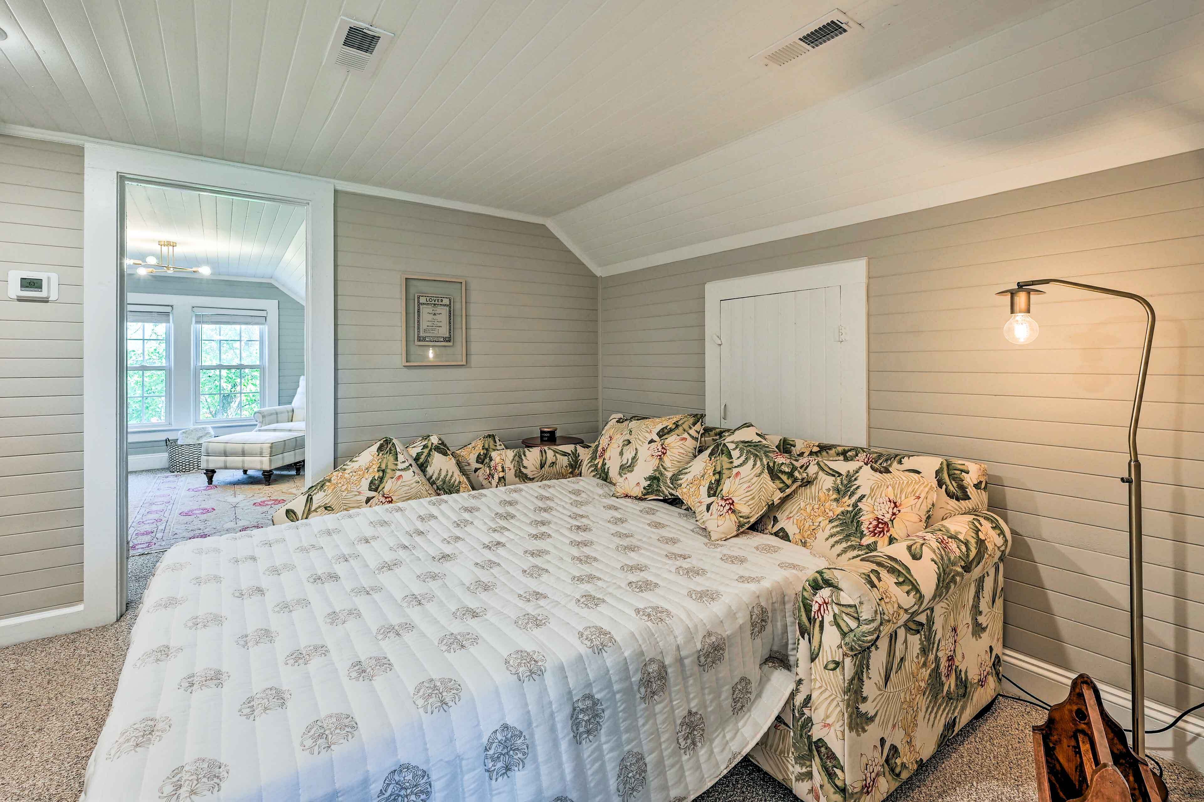 A bedroom with a large bed, floral-patterned sofa, a floor lamp, and a view of a porch.