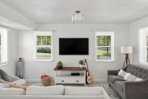 A living room with a flat-screen TV, two sofas, a coffee table, a guitar, a lamp, and a large window with blinds. - Meadow Retreat' Near Parks & USU (Logan)