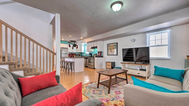 Un salon spacieux avec un canapé gris, des coussins rouges, un canapé beige avec des coussins turquoise, une table basse en bois et un tapis coloré. On aperçoit au fond une cuisine avec des meubles blancs et un coin repas équipé d’un téléviseur et de tabourets de bar.