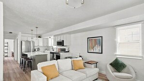 A modern living room with a sofa, a coffee table, and a chair. There is a kitchen area with cabinets and appliances in the background. - Meadow Retreat' Near Parks & USU (Logan)