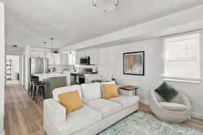 A modern living room with a sofa, a coffee table, and a chair. There is a kitchen area with cabinets and appliances in the background. - Meadow Retreat' Near Parks & USU (Logan)