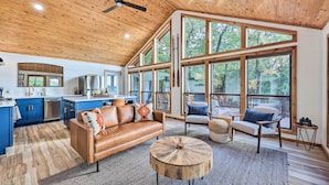 A spacious living area with a wooden ceiling, large windows, a leather sofa, and a round coffee table. - 'foxtail' Cabin Retreat: Steps to Beaver Lake! (Rogers)