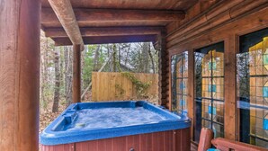 A wooden cabin with a hot tub under a covered area, surrounded by a wooden fence and trees.