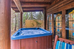Une cabane en bois avec un bain à remous situé sous une zone abritée, entourée d’une clôture en bois et d’arbres.