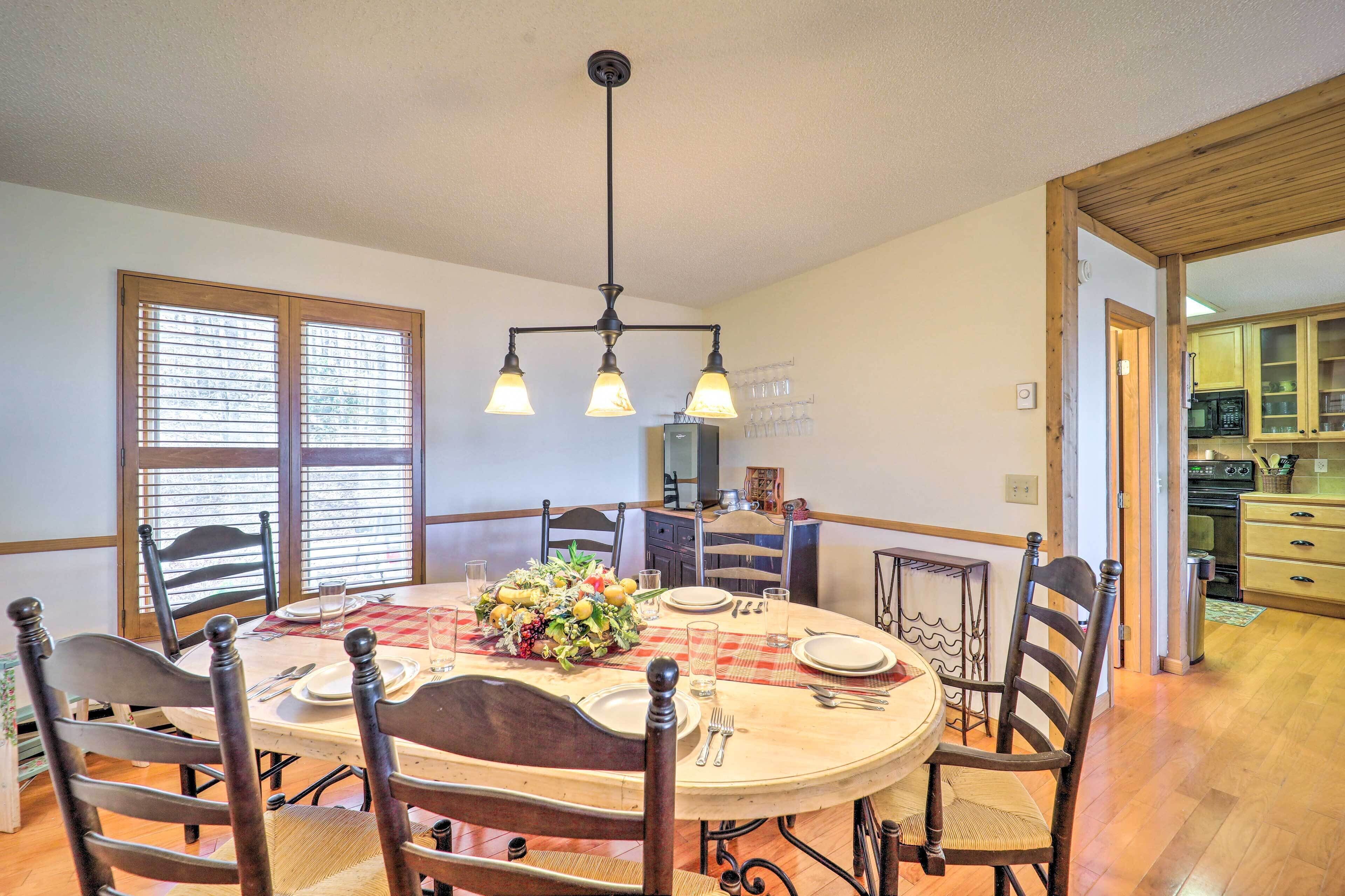 Un comedor con una mesa redonda dispuesta para seis personas, rodeada de sillas de madera. Un candelabro cuelga sobre la mesa, y se ve una ventana con persianas en el fondo.