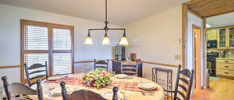 Un comedor con una mesa redonda dispuesta para seis personas, rodeada de sillas de madera. Un candelabro cuelga sobre la mesa, y se ve una ventana con persianas en el fondo.