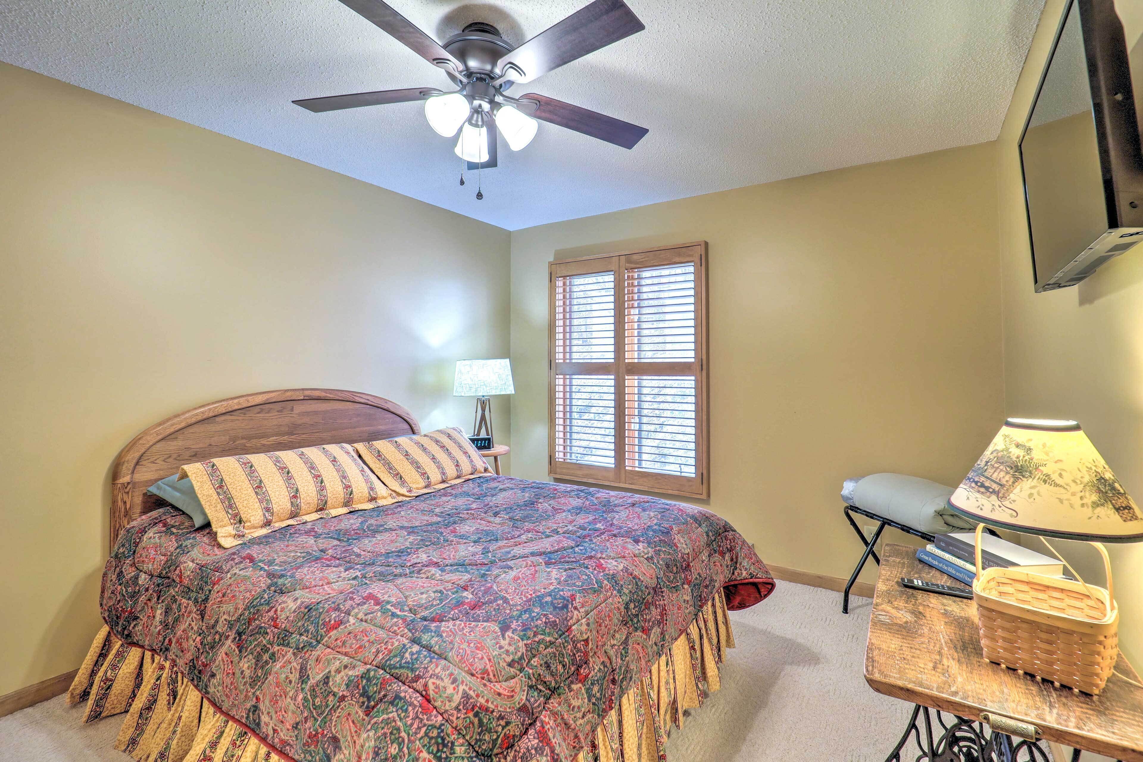 A bedroom with a bed, a ceiling fan, a window with blinds, a nightstand with a lamp, and a table with a basket.