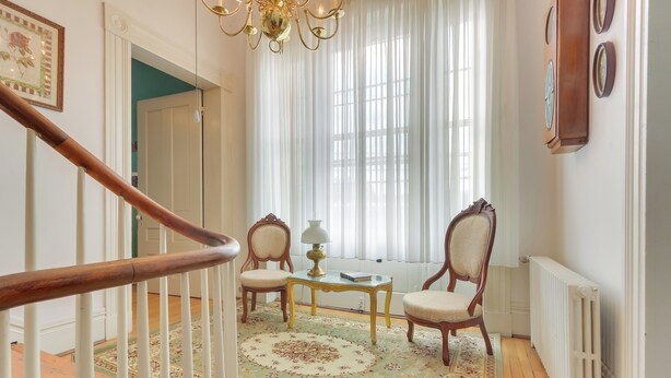 A staircase with a wooden handrail, a patterned rug, two wooden chairs, a table with a lamp, and a radiator on the wall. - Historic West Virginia Home Built in 1854! (Moorefield)