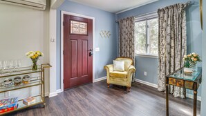 A room with a wooden floor, a red door, a vintage armchair, a glass coffee table, and a wall-mounted shelf with decorative items. - 'house in the Woods' in Ooltewah w/ Fire Pit! (Ooltewah)