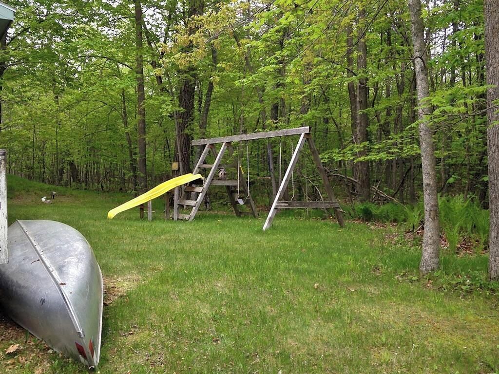 A wooden swing set in a wooded area with a metal boat nearby.