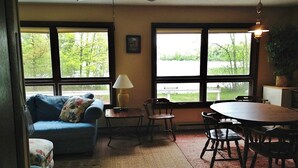 Un salon comprenant un canapé, des fauteuils, une table et une lampe. De grandes fenêtres offrent une vue sur les arbres et une pelouse.