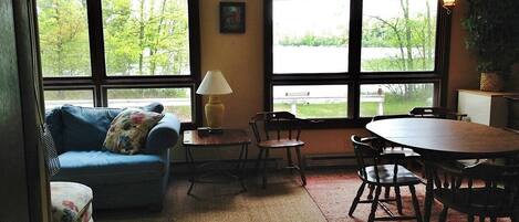 Una sala de estar con un sofá, sillas, una mesa y una lámpara. Amplias ventanas ofrecen vistas a árboles y un jardín.