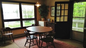 Un coin repas avec une table ronde et six chaises, de grandes fenêtres donnant sur un paysage verdoyant, une plante suspendue et une porte à motifs géométriques.