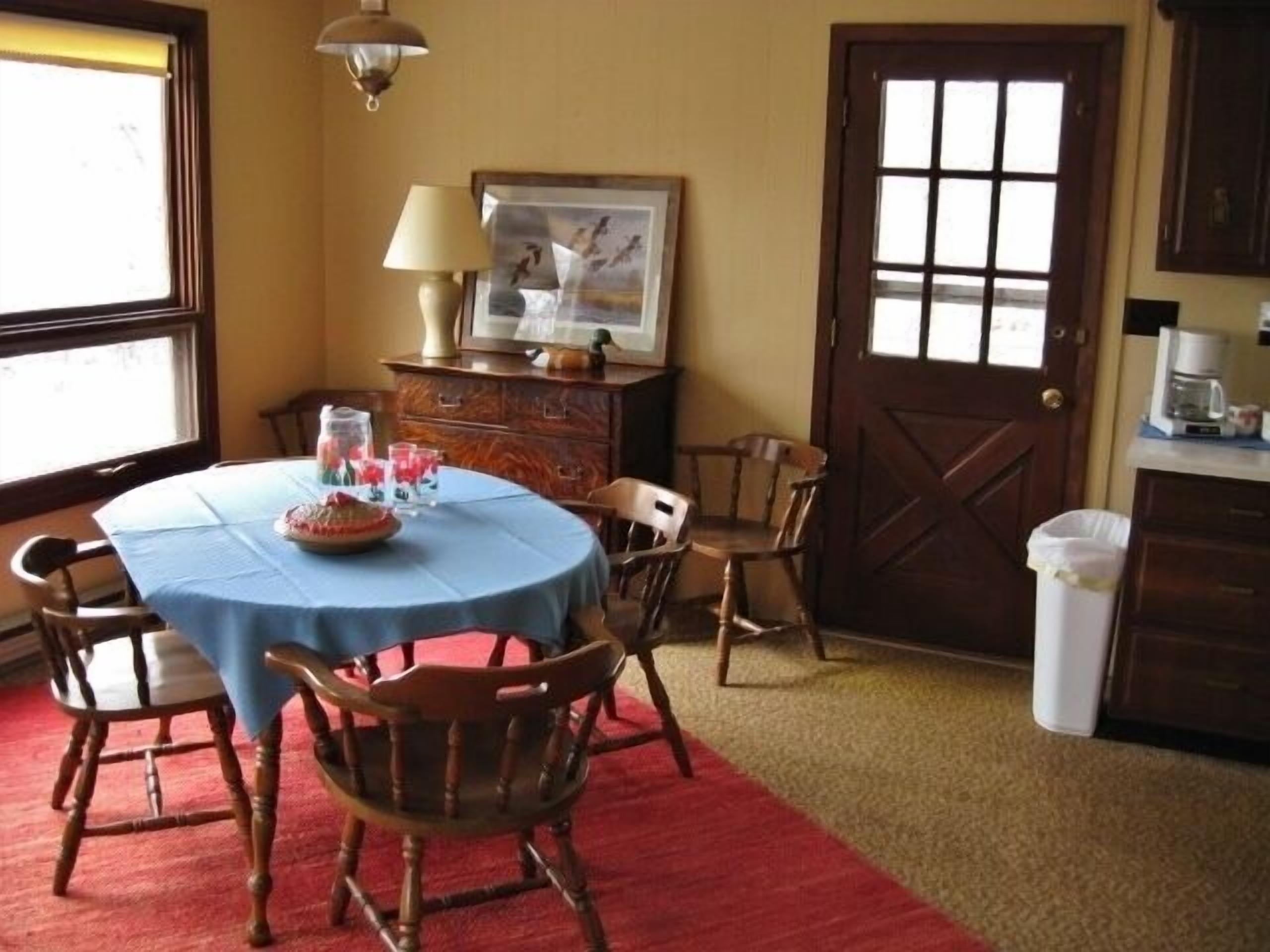 A dining area with a round table covered with a blue tablecloth, surrounded by wooden chairs. There is a wooden cabinet with a lamp and a framed picture on top. A door with glass panels is visible in the background.