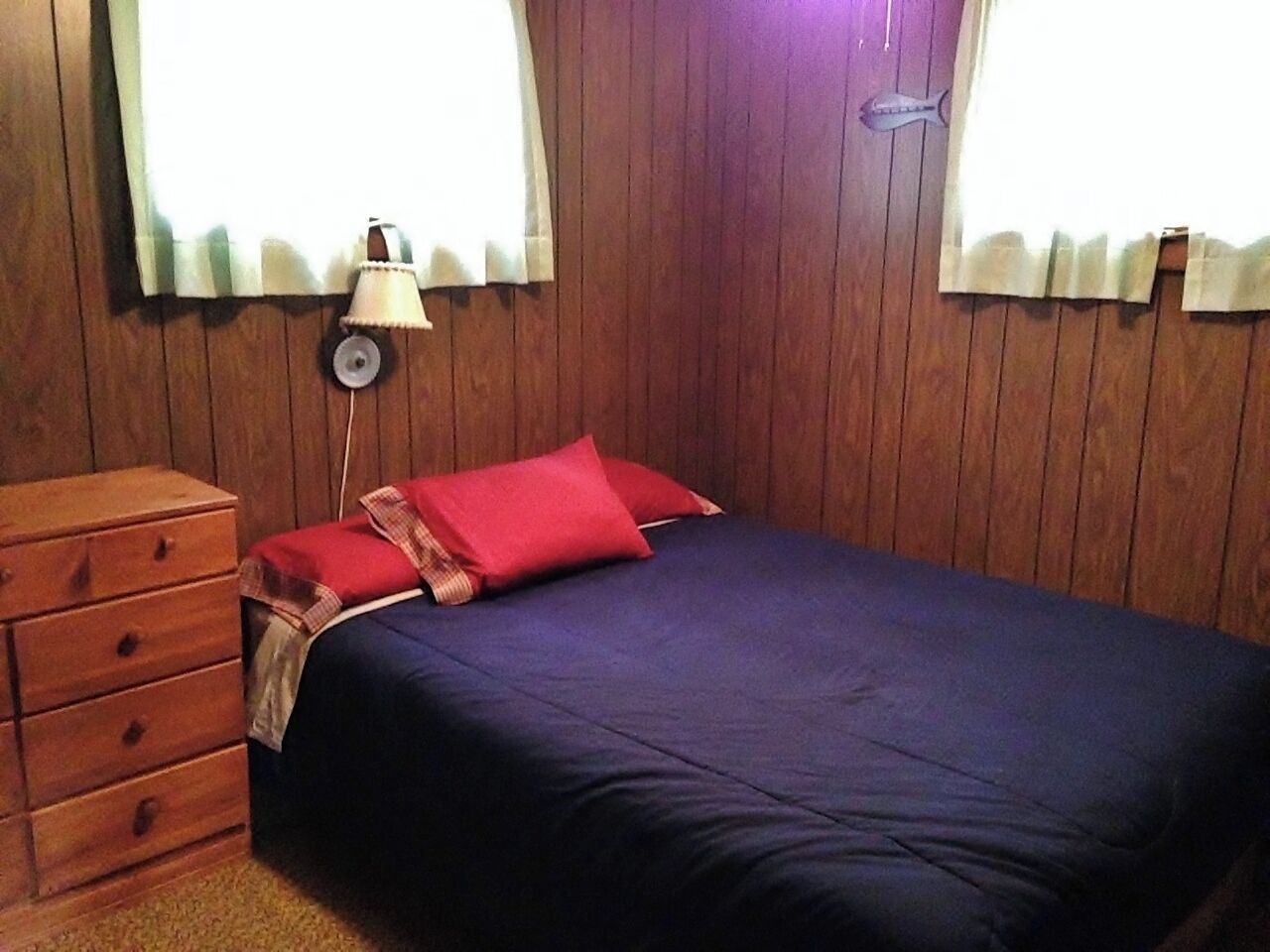 Une chambre avec un mur en bois, un lit recouvert d’une couvre-lit bleu foncé et agrémenté d’oreillers rouges, une commode en bois et une lampe posée sur la commode.