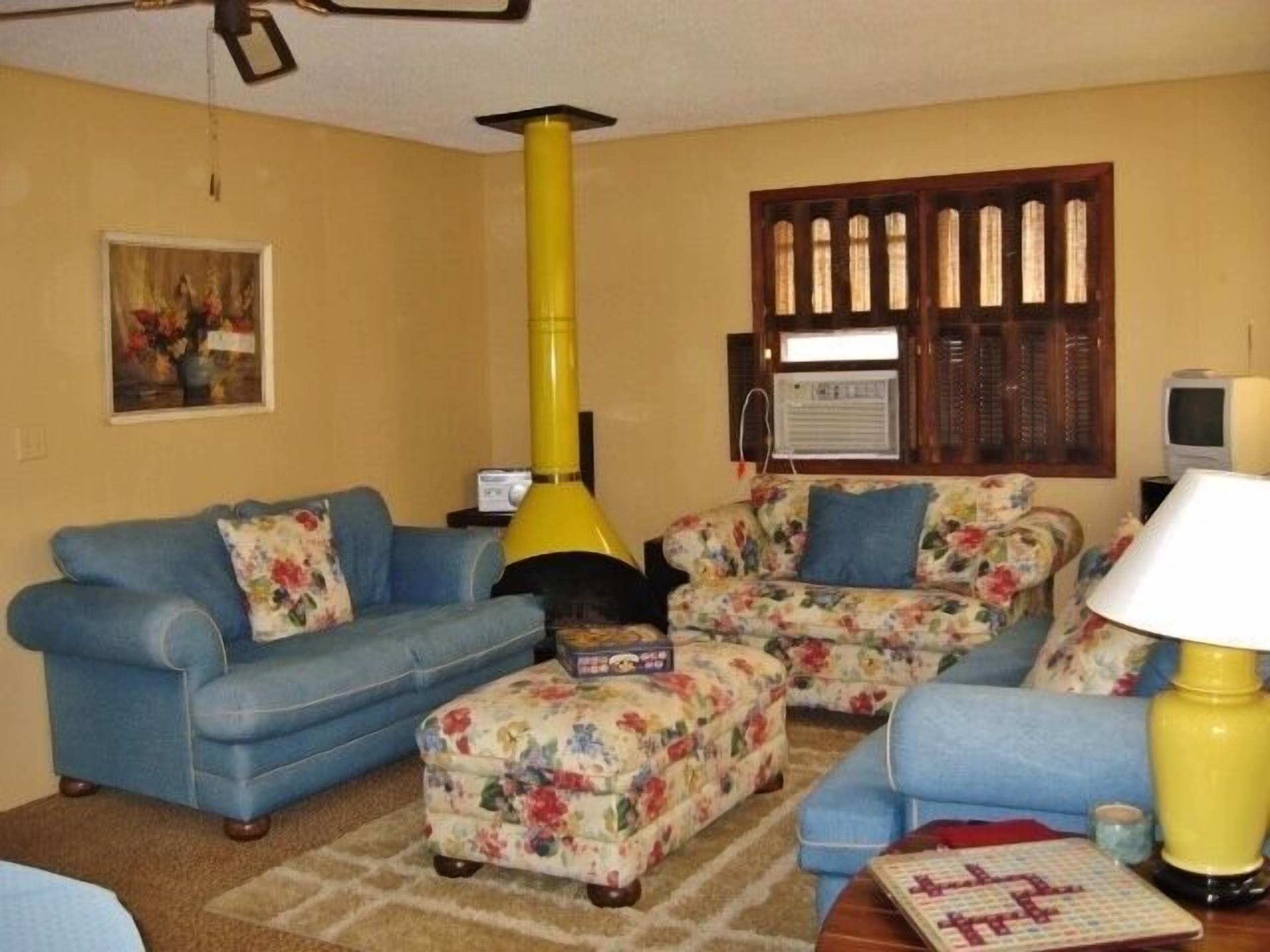 A living room with a blue sofa, floral ottoman, yellow lamp, and a television.