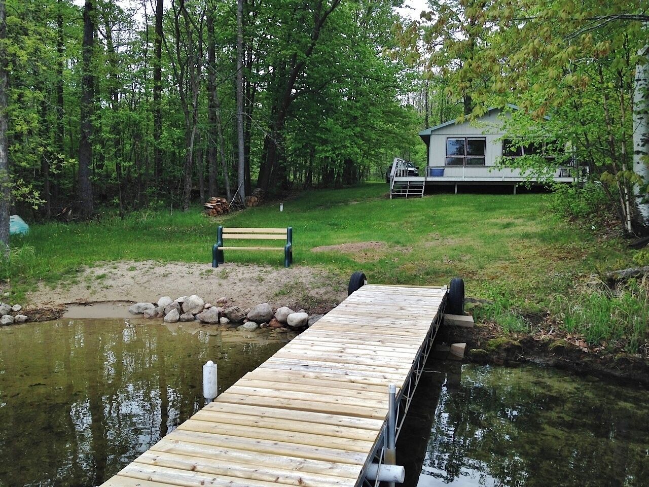 A wooden dock extending into a pond, surrounded by trees and a grassy area with a bench and a house in the background.