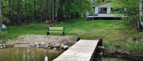 A wooden dock extending into a pond, surrounded by trees and a grassy area with a bench and a house in the background.