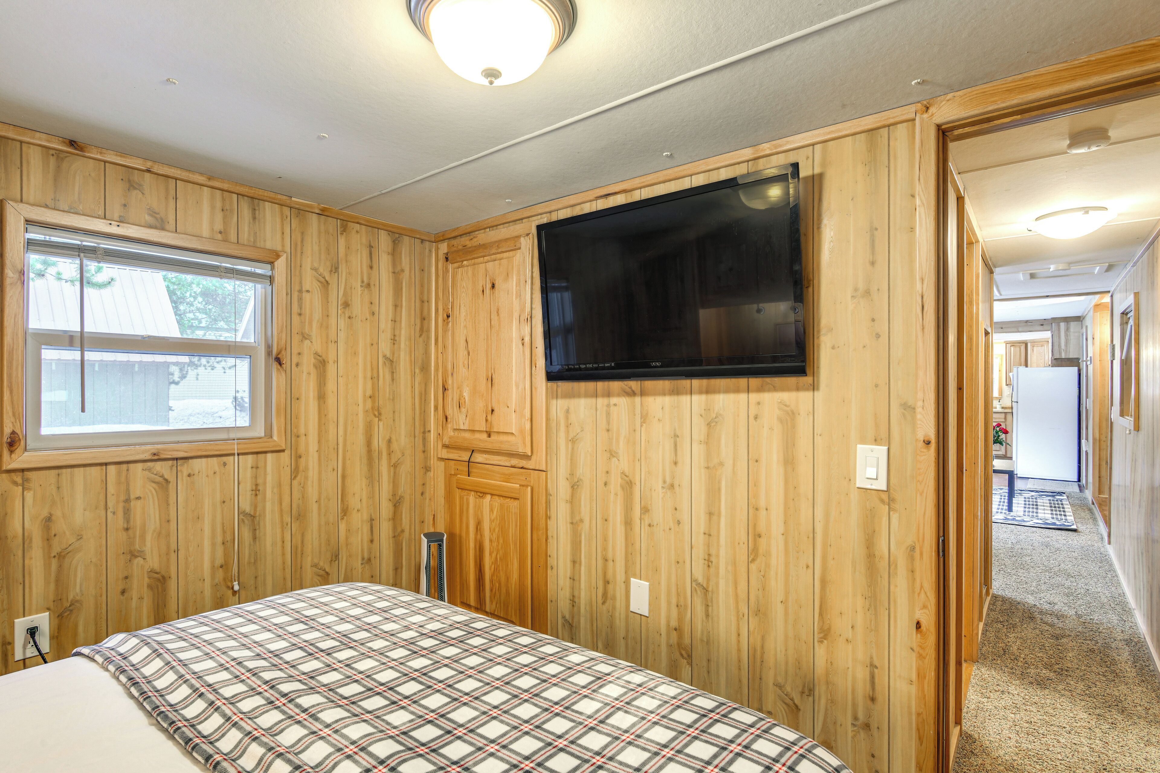 A bedroom with wooden walls, a large window, a bed with a plaid bedspread, and a flat-screen TV mounted on the wall.