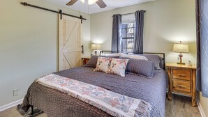 A bedroom with a bed, a wooden nightstand, a ceiling fan, a window with curtains, and a door with a barn-style design. - Classic Lake O' the Pines Retreat! Dock, Fire Pit (Jefferson)