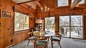 A wooden interior with a round table, chairs, a chandelier, and large windows.