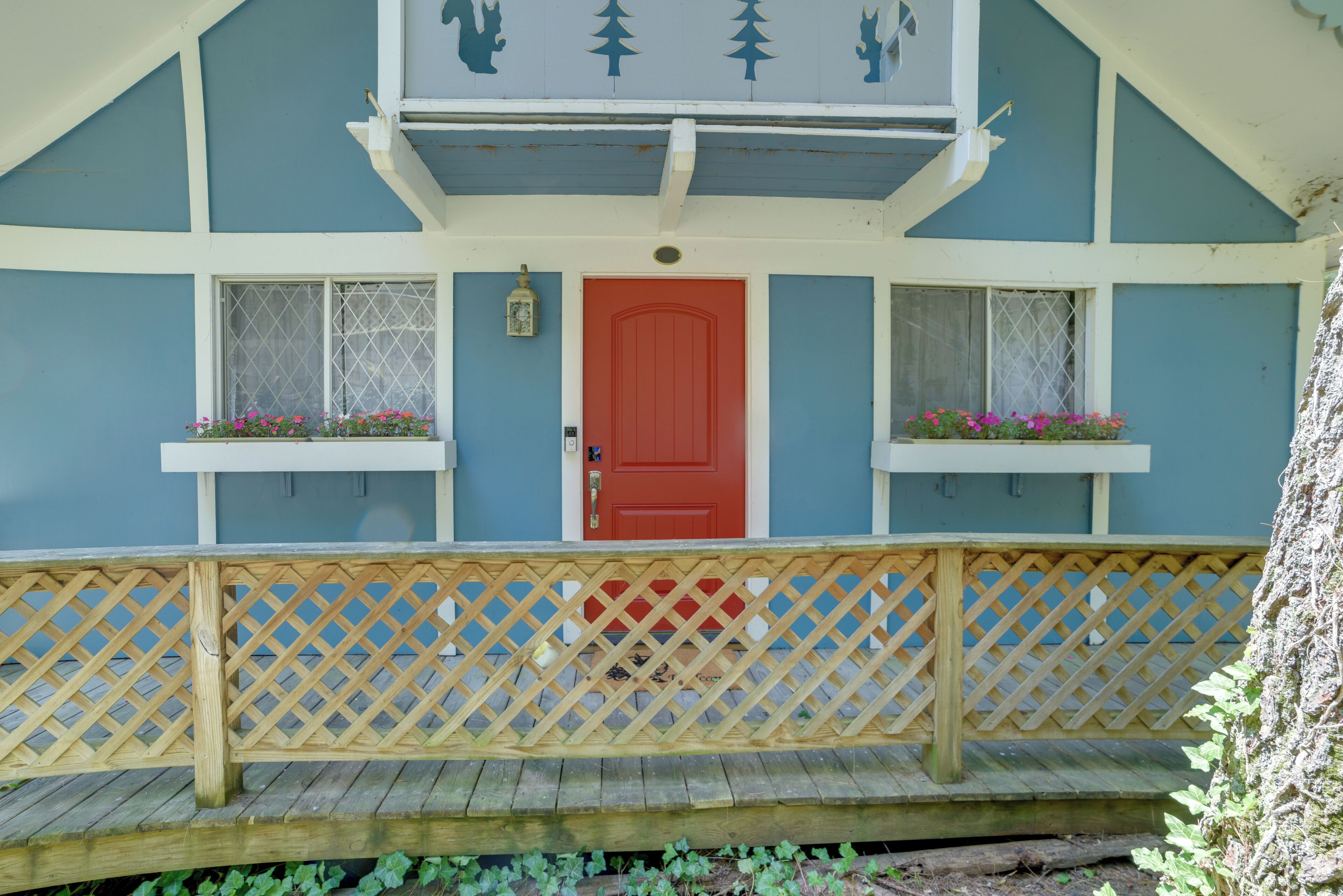 Une terrasse en bois avec une balustrade en treillis devant un bâtiment doté d’une porte rouge et de deux fenêtres ornées de jardinières.
