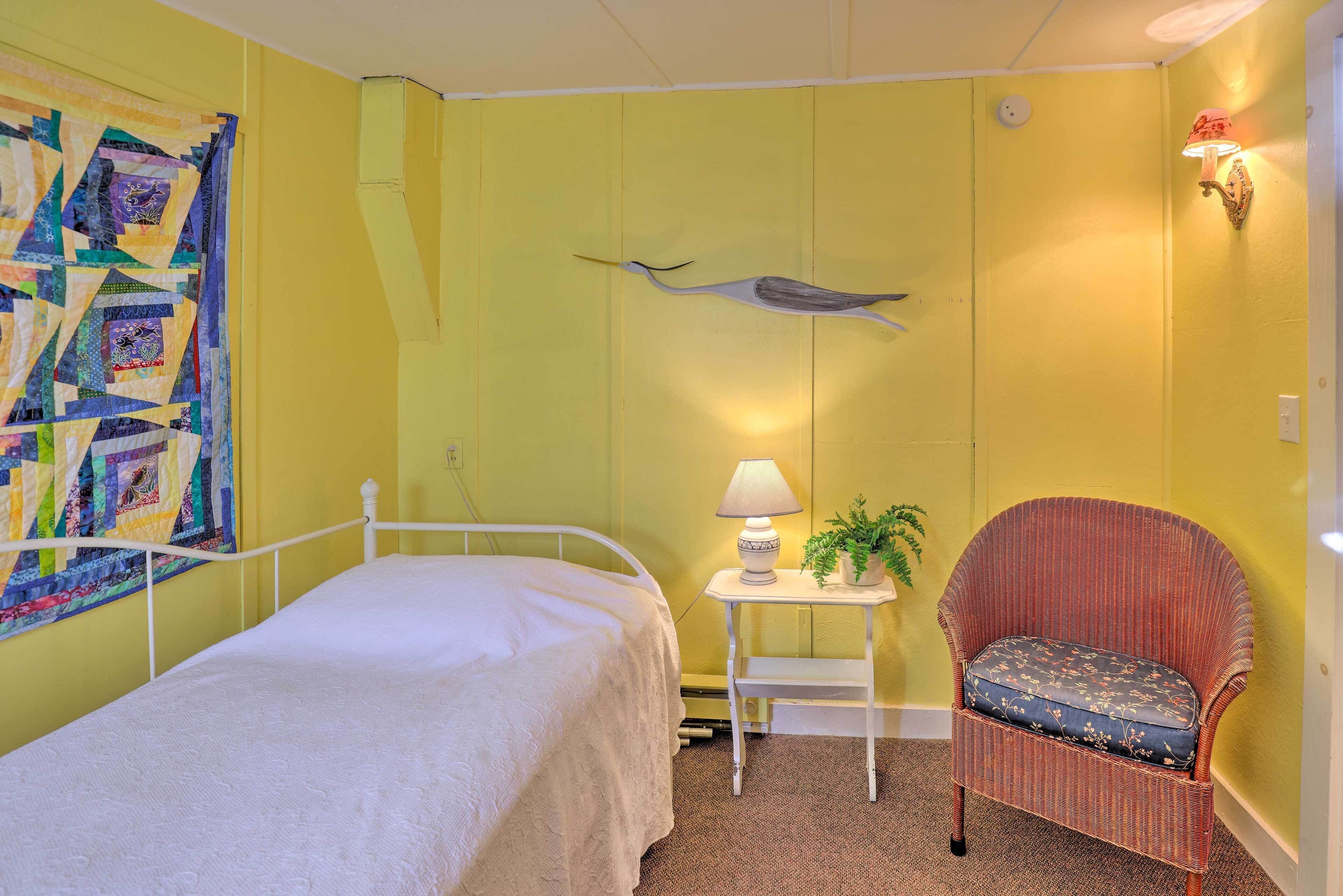 A room with a bed, a chair, a small table with a lamp, and a wall hanging.